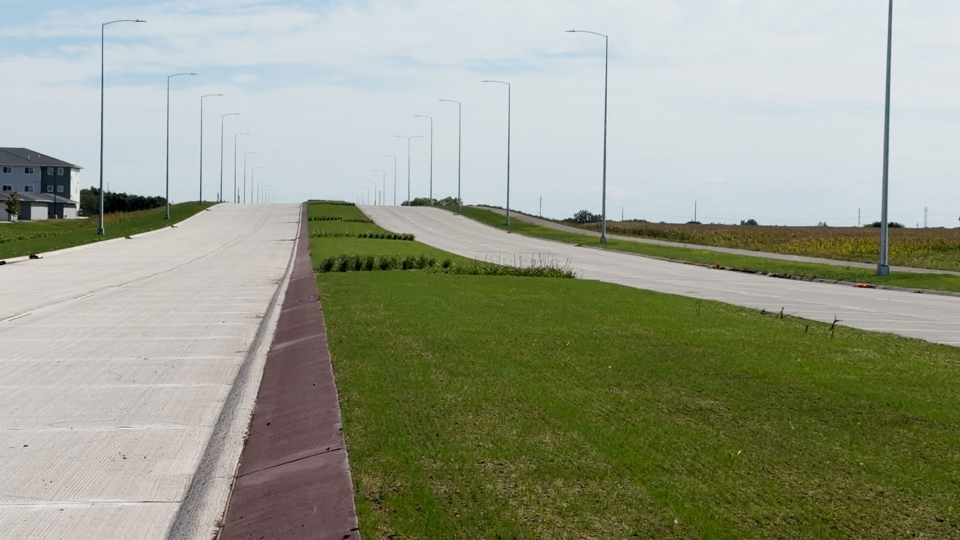 South Veterans Parkway southbound stretches over a gentle rise, with concrete pavement, landscaped median, streetlights, and grassy shoulders extending into the distance. Agricultural fields and residential buildings are visible along the corridor.