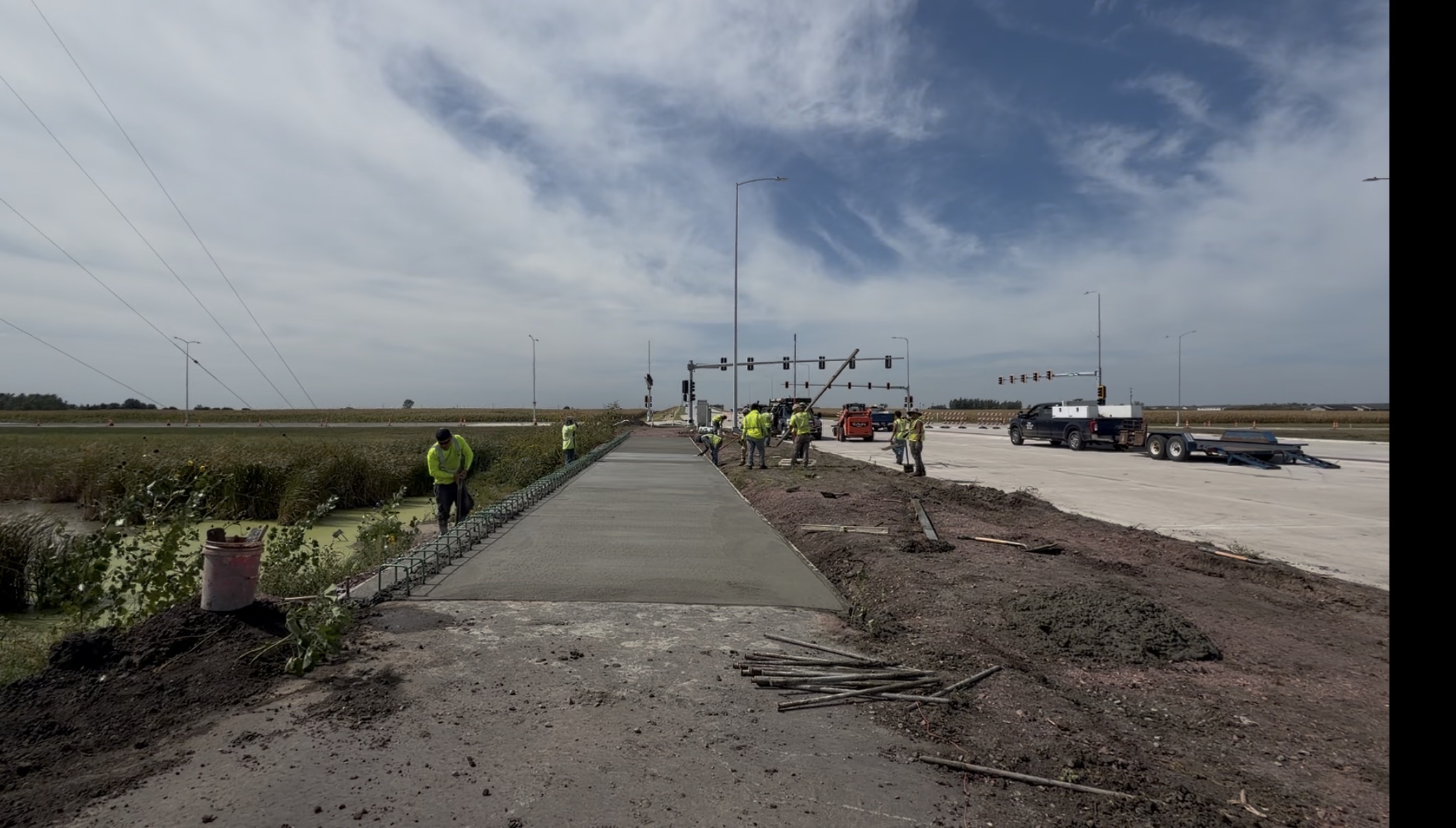 Road construction crew works on a newly poured concrete sidewalk or trail near Cliff Avenue intersection, with construction vehicles, traffic signals, and wetlands visible in the background under a partly cloudy sky.