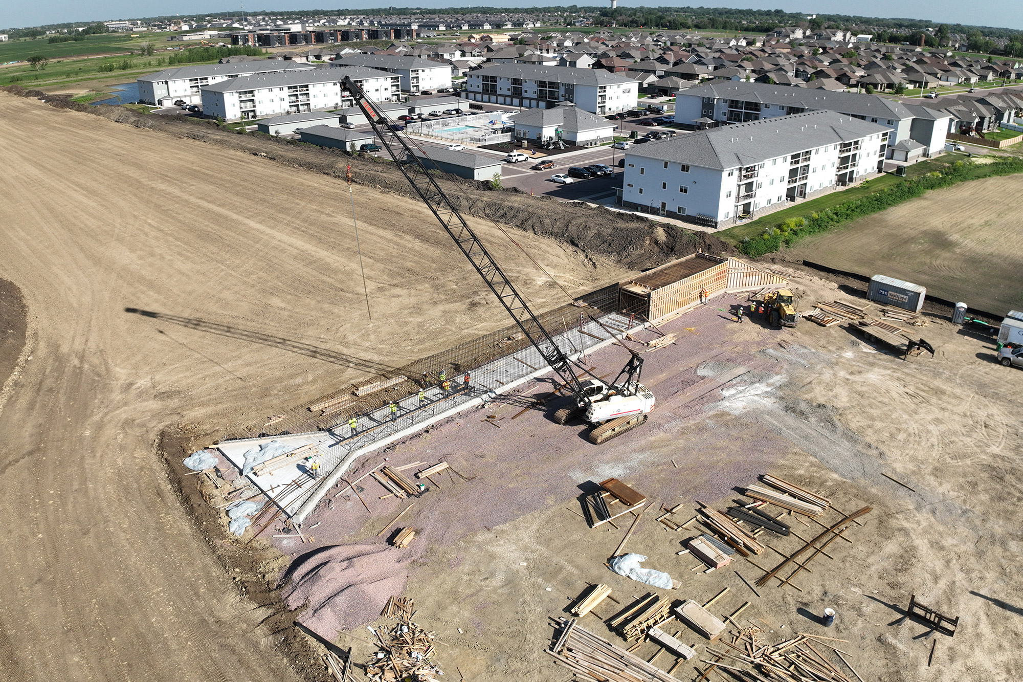 Drone view of active construction. A crane is moving material into place. Bulk materials visible.